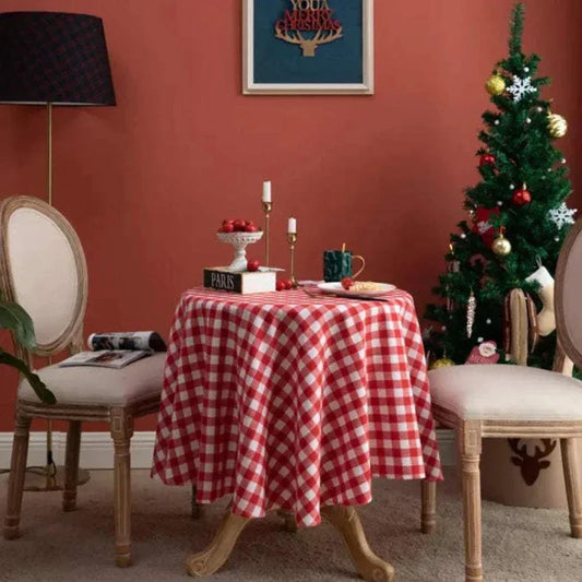 Round red and white plaid Christmas tablecloth on dining table with chairs and decorated Christmas tree in background