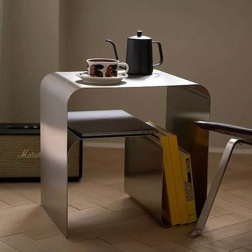 Minimalist stainless steel metal side table with curved edges holding a teapot, cup, and books in a modern living room setting