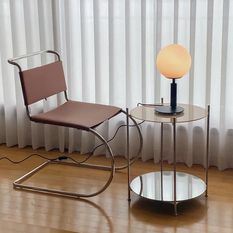 Minimalist stainless steel dining chair with modern design, armless and leather seat, next to a round table with lamp.