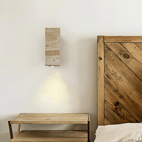 Elegant travertine stone wall light with warm LED glow mounted above wooden bedside table in bedroom
