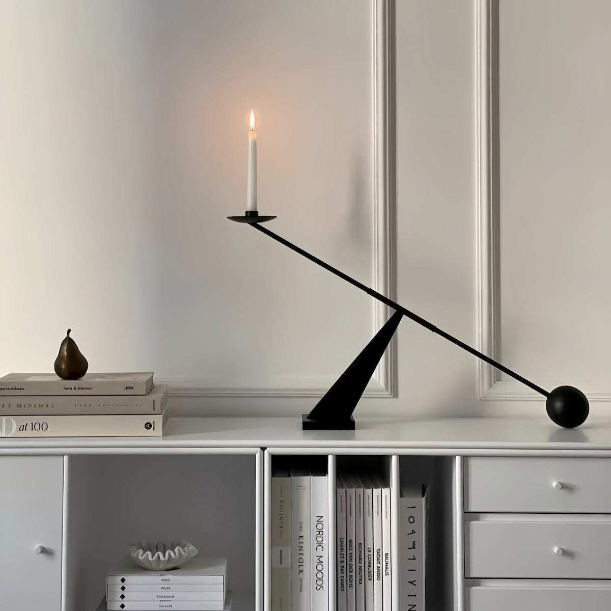 Black minimalist geometric metal candle holder with a lit white taper candle on a white cabinet against a plain wall