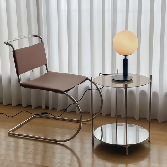 Minimalist stainless steel dining chair with brown leather seat and backrest, modern armless design next to glass and metal side table with round lamp