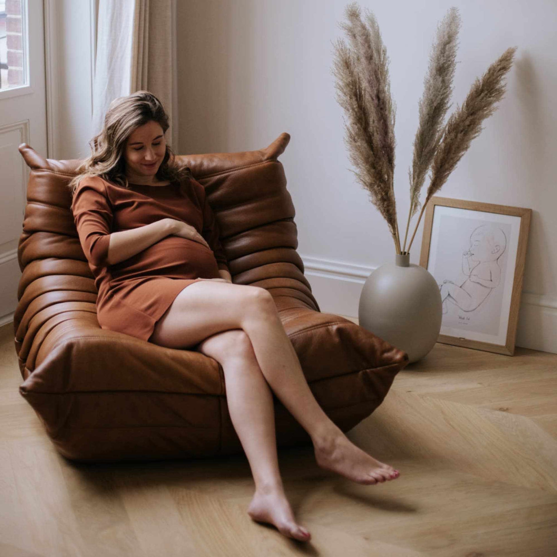 Woman relaxing on brown leather Togo Chair in modern living room with pampas grass and framed artwork